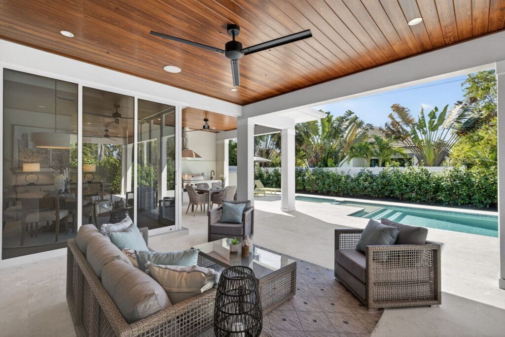 A bright, professionally staged covered patio featuring a gray wicker sofa and armchairs arranged around a glass coffee table. The space features a warm wood-plank ceiling with a black fan and opens up to a beautifully presented backyard swimming pool framed by tropical greenery.