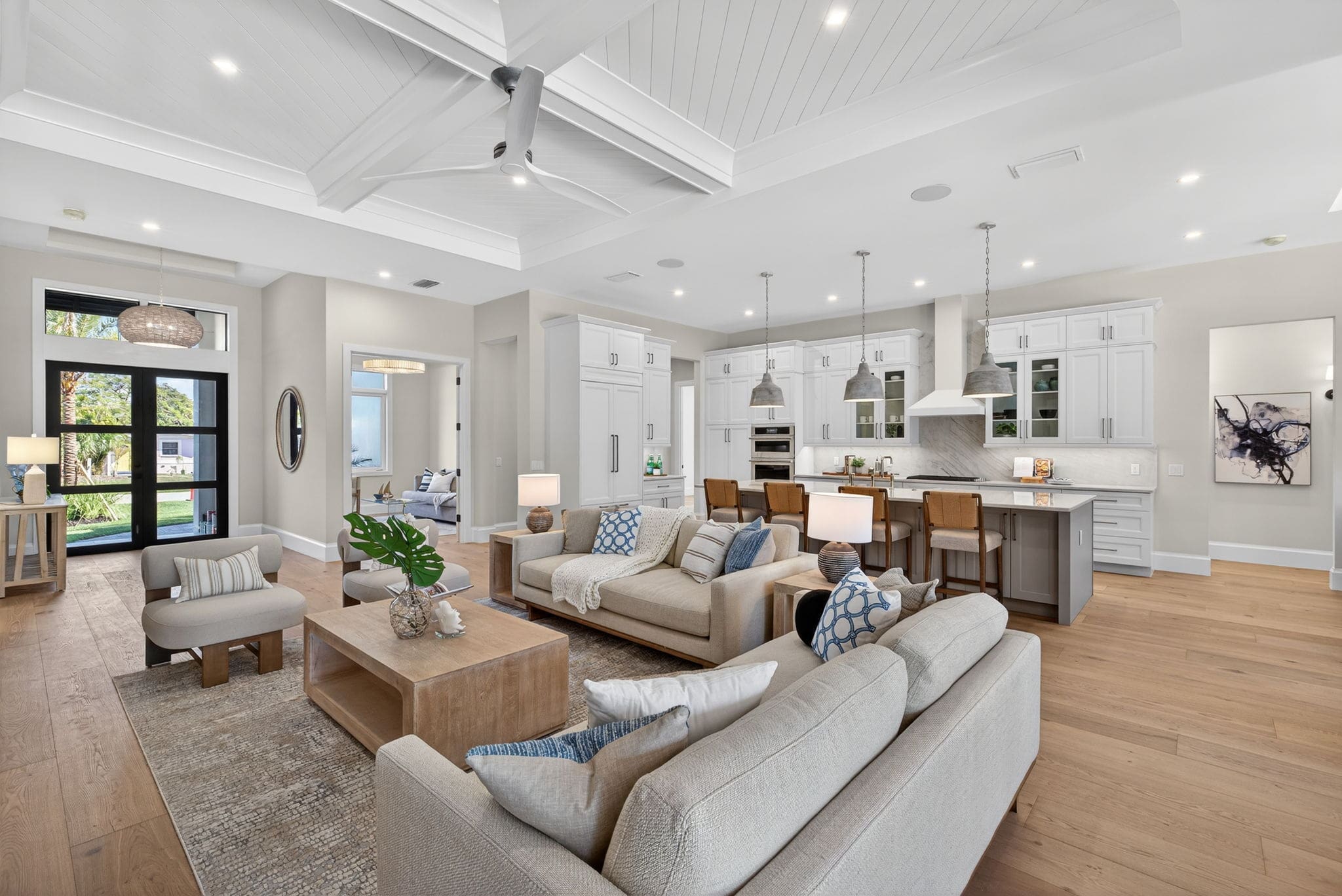 A bright, professionally staged open-concept living room and kitchen with modern coastal decor. The living area features light beige sofas, blue and white patterned accent pillows, and a wooden coffee table on a textured rug. The spacious white kitchen in the background includes a large island with barstools and industrial pendant lights.