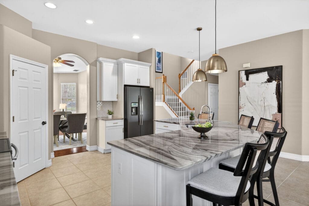 A cohesive kitchen staged by stageandamaze, showing a white island with a marble countertop, black stools, and neutral tones that create a unified, high-end look.