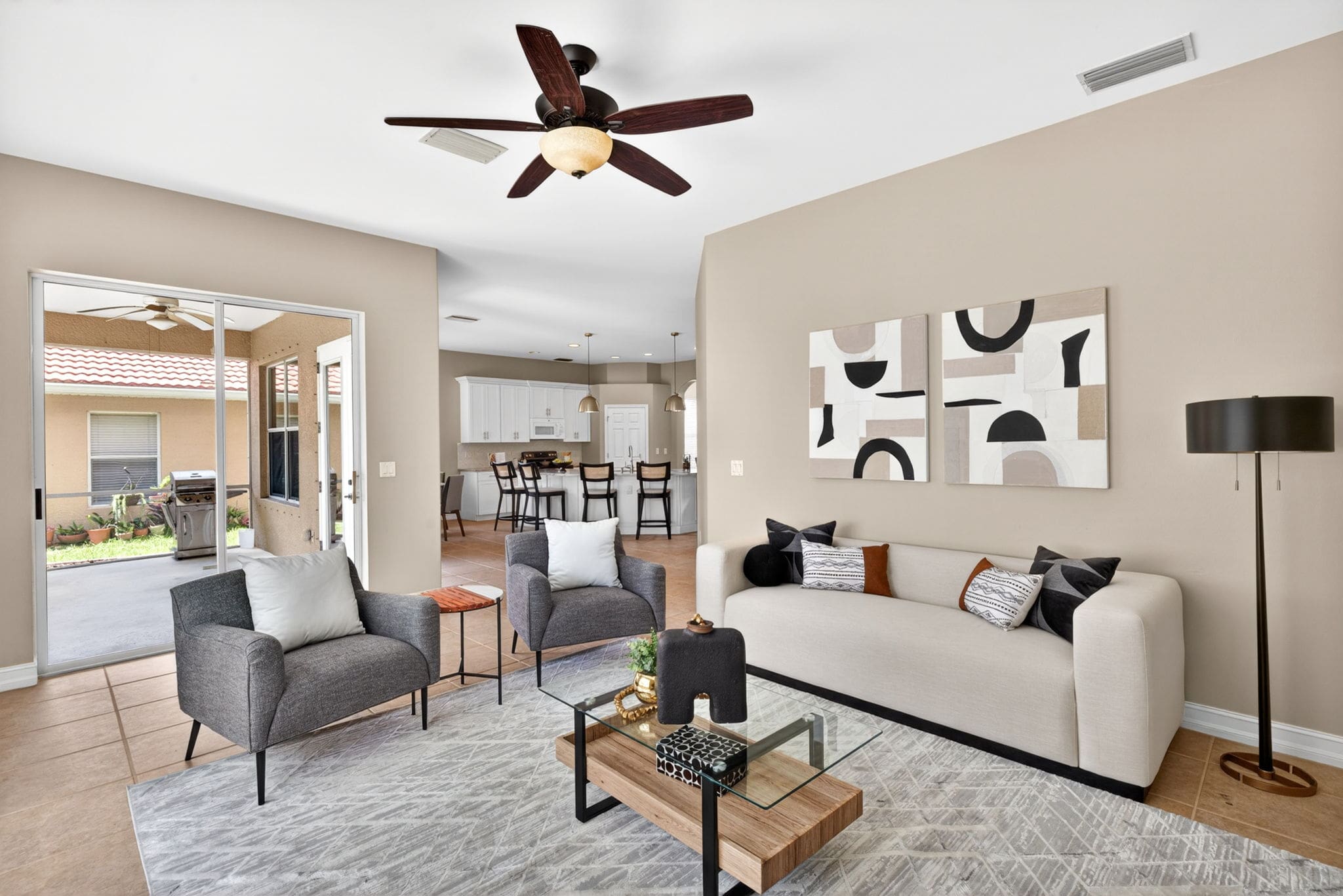 A modern, open-concept living room staged by stage and amaze, featuring a beige sofa, grey armchairs, a glass coffee table, and abstract wall art, opening into a white kitchen.